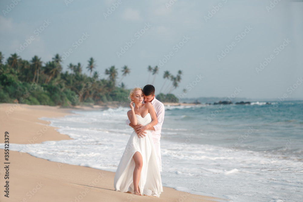 Newlyweds sharing a romantic and fun moment at the beach. Groom hug the bride from the back and kiss in the cheek, walkig barefoot tropical beach Newlyweds sharing a romantic and fun moment at the beach. Groom hug the bride from the back and kiss in the cheek, walkig barefoot tropical beach