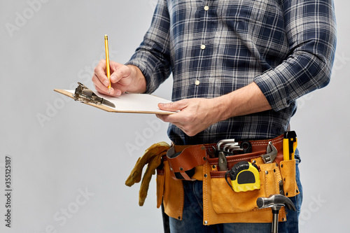 repair, construction and building - male worker or builder with clipboard, pencil and working tools on belt over grey background