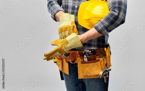 repair, construction and building - male worker or builder with helmet and working tools on belt putting protective gloves on over grey background © Syda Productions