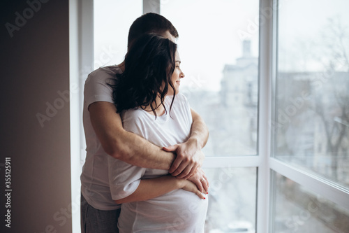 Pregnant couple cuddling each other at home. Man holding pregnant woman's stomach in bedroom. Pregnant young woman are waiting for a child at home by the window. Loving family, husband and wife.