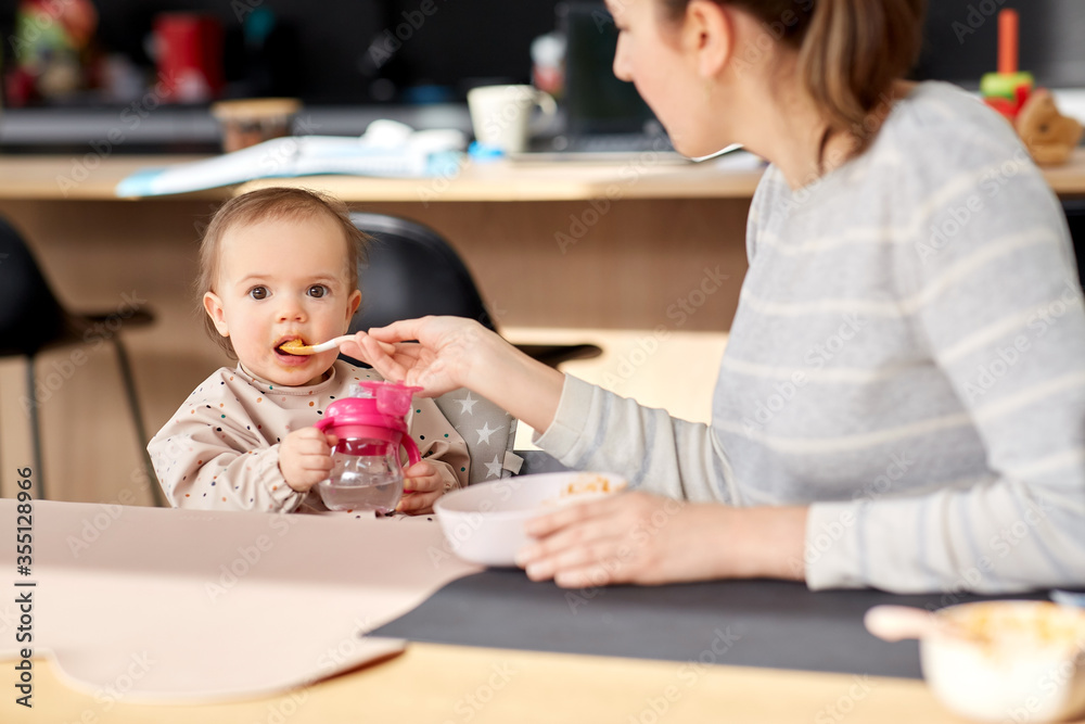 family, eating and people concept - happy mother with puree and spoon feeding little baby girl sitting in highchair at home