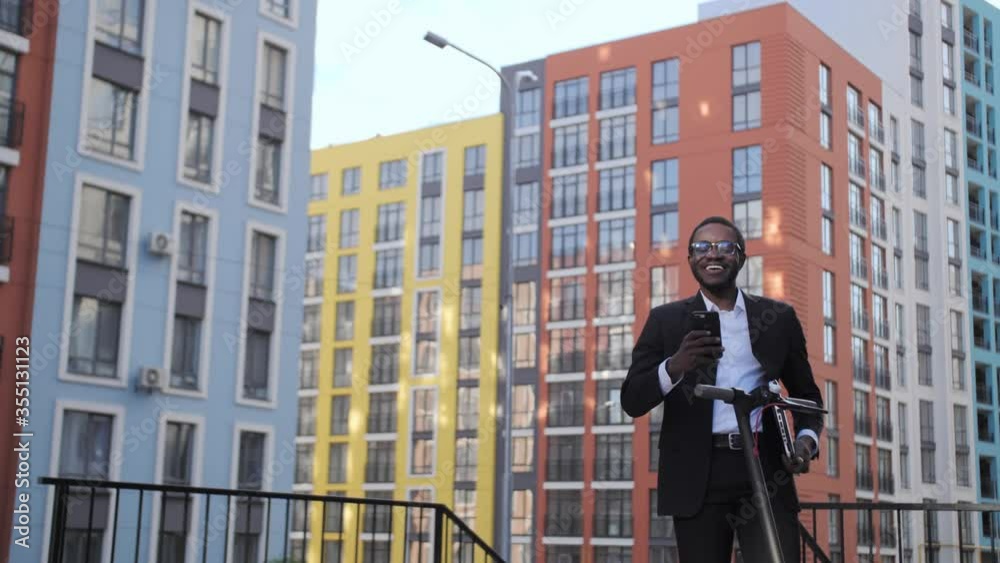 Handsome dark skinned businessman in a black suit make video call on mobile outdoors on the background of a colored houses, next to him is an electric scooter. Handsome dark skinned businessman in a black suit make video call on mobile outdoors on the background of a colored houses, next to him is an electric scooter.