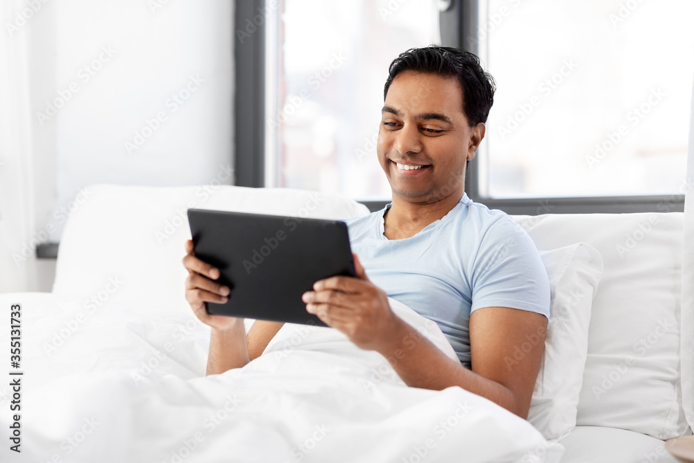 people, bedtime and rest concept - happy smiling indian man with tablet pc computer lying in bed at home people, bedtime and rest concept - happy smiling indian man with tablet pc computer lying in bed at home