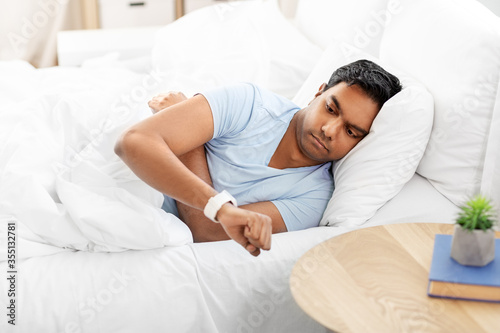 people, sleeping and rest concept - indian man looking at smart watch in bed at home