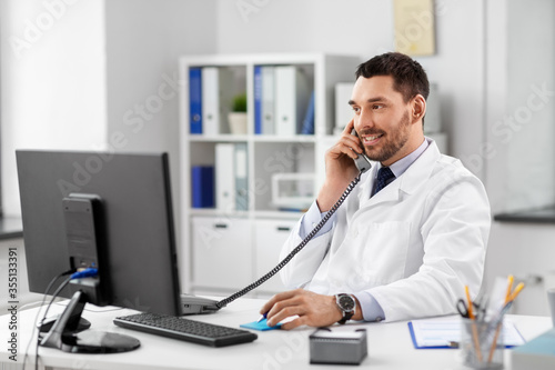healthcare, medicine and people concept - smiling male doctor with clipboard calling on desk phone at hospital