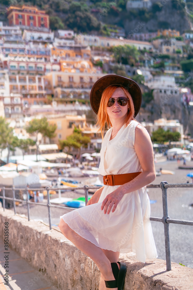Fototapeta premium A girl in a white dress, sunglasses and a hat stands on the beach in Positano. View of houses and hotels in the background. Travel and vacation. Copy space place for text. Vertical photo