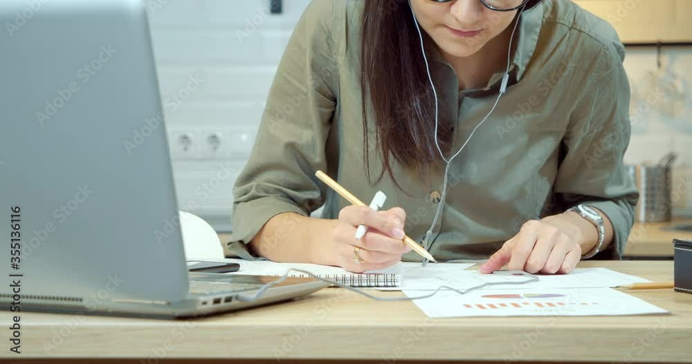 Close-up of a young woman checking a project drawing and writing down data in a notebook at the workplace.