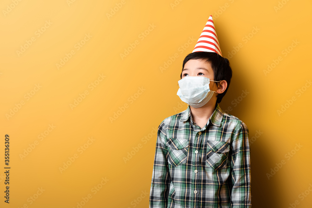 Obraz premium asian boy in birthday party cone and medical mask on yellow during self isolation