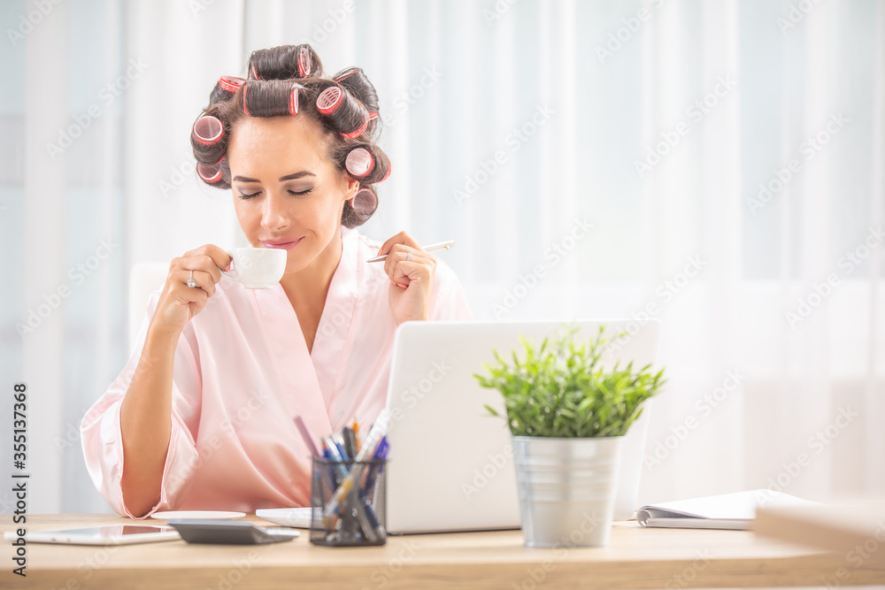 Woman with hair rollers and nighgown smells coffee while sitting in front of computer at home Woman with hair rollers and nighgown smells coffee while sitting in front of computer at home
