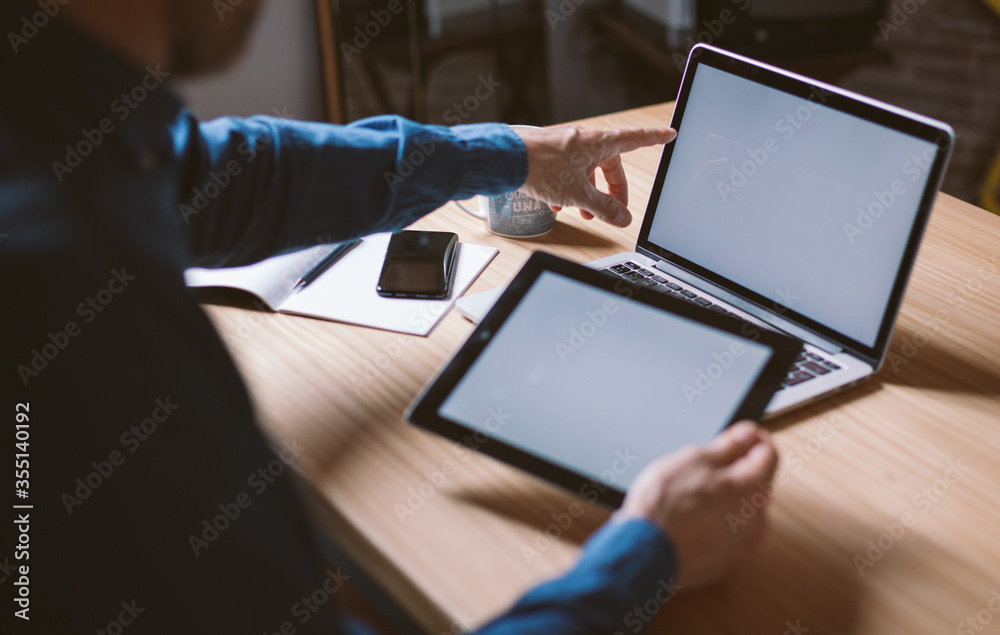 Male hand pointing at laptop white screen holding a tablet. Mockup of laptop and tablet screen. Modern organic wooden environment Male hand pointing at laptop white screen holding a tablet. Mockup of laptop and tablet screen. Modern organic wooden environment