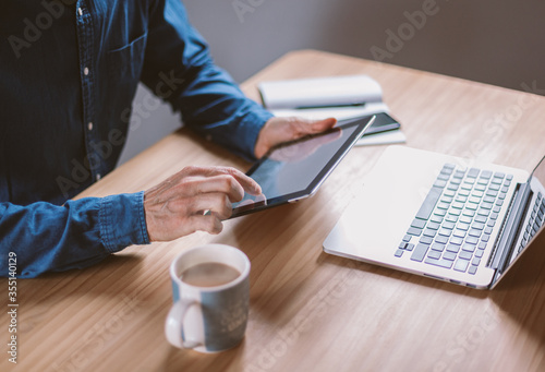 Closeup on male hands working on his tablet and laptop on wooden table having tea. Businessman working remotely from cosy home or coworking place.