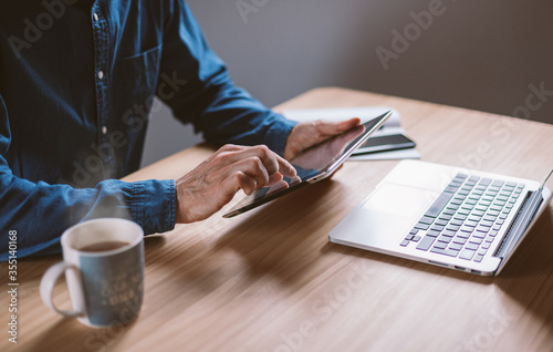 Closeup on male hands working on his tablet and laptop on wooden table having tea. Businessman working remotely from cosy home or coworking place.