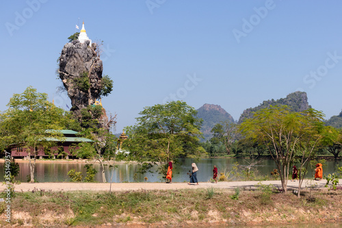 Buddhist monks walking dressed in orange, passing in front of Kyaut Ka Latt pagoda. Buddhist monastery and the sacred rock surrounded by a lake. Hpa An, Myanmar, Burma, Southeast Asia photo