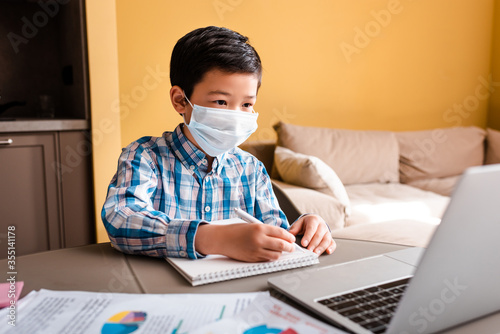 asian boy in medical mask writing and studying online with laptop at home during quarantine