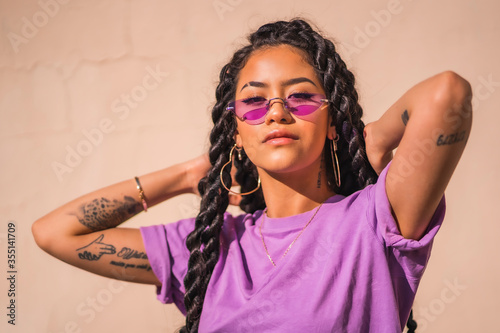 Urban session. Young dark-skinned woman with long braids wearing purple glasses on a plain cream-colored background, with serious look