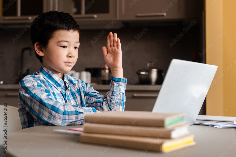 asian child with hand up studying online with laptop at home during self isolation asian child with hand up studying online with laptop at home during self isolation