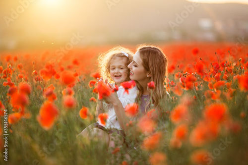 Beautiful smiling baby girl with mother are having fun in field of red poppy flowers over sunset lights, spring time © _chupacabra_