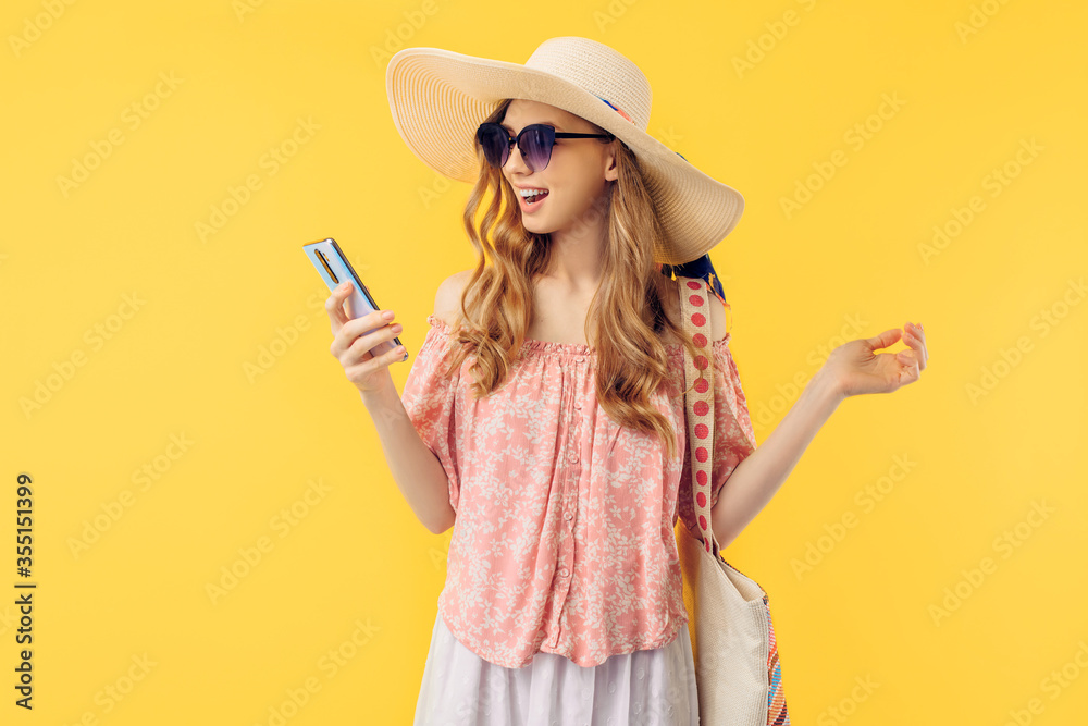smiling young woman in a summer hat and sunglasses, holding a smartphone on an yellow background smiling young woman in a summer hat and sunglasses, holding a smartphone on an yellow background