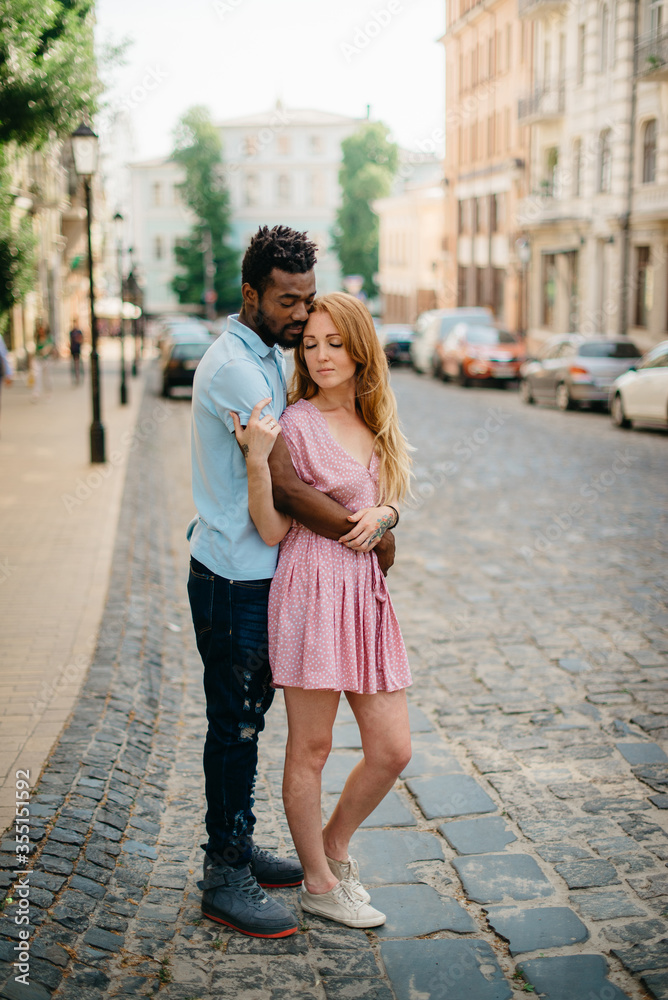 Interracial Relations. An African man and a Caucasian woman stand embracing on a city street. Full height portrait.