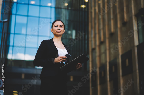 Successful executive manager dressed in elegant outfit holding modern telephone and folder in hands standing outdoors against office promotional background.Prosperous banker with digital cellular