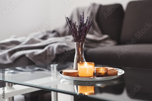 Coffee table design idea: aroma candles and dried lavender bouquet on a metal tray, sofa with grey blanket on background. Simple scandinavian home decor. Hygge concept