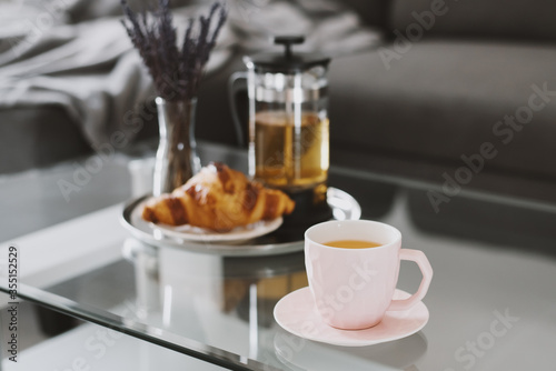 Herbal tea in a pink cup, fresh croissant, french press tea pot and dried lavender bouquet served on a coffee table in living room. Breakfast concept