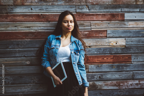 Half length portrait of confident young woman with long hair holding modern tablet with mock up area and looking at camera.Charming female dressed in denim jacket standing on promotional background