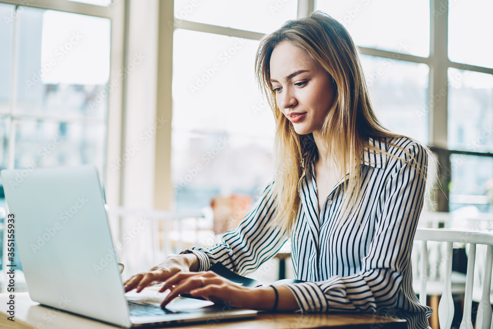 Fototapeta premium Cheerful female copywriter keyboarding text on digital netbook and searching information on internet websites for productive work.Positive freelancer updating software on laptop device sitting indoors
