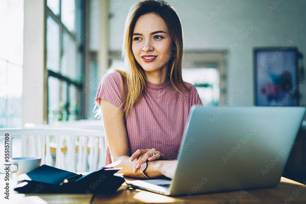 Attractive smiling businesswoman sending messages via laptop computer while looking away and thinking on research for design startup project working remotely in stylish coffee shop interior Attractive smiling businesswoman sending messages via laptop computer while looking away and thinking on research for design startup project working remotely in stylish coffee shop interior