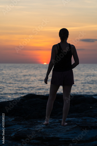 Young woman standing on some rocks on the coast enjoying the sunset. She is barefoot and wearing shorts and a tank top. In the background you can see the sea and the sun setting over the horizon.