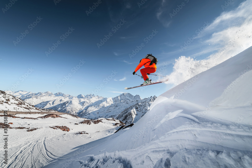 An athlete male skier jumps from a snow-covered slope against the backdrop of a mountain landscape of snow-covered mountains on a sunny day. The concept of winter sports