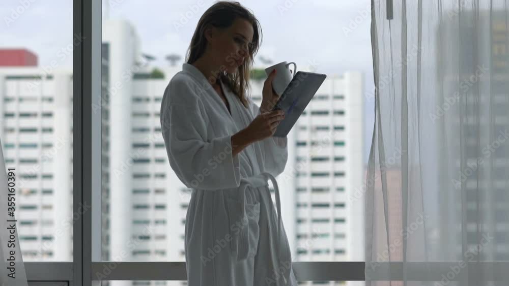 Young woman in bathrobe with tablet computer drinking coffee by the window at home Young woman in bathrobe with tablet computer drinking coffee by the window at home