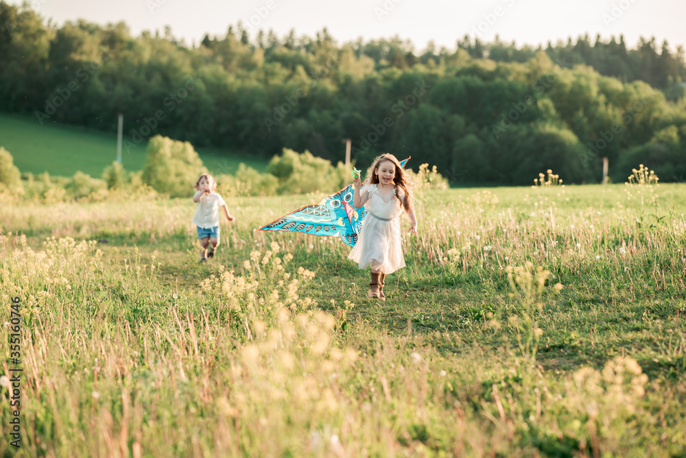 Fototapeta premium Cute curly little girl in white dress and boy in white shirt, running on sunset meadow with kite. Healthy, enjoy countryside summer and happy childhood concept