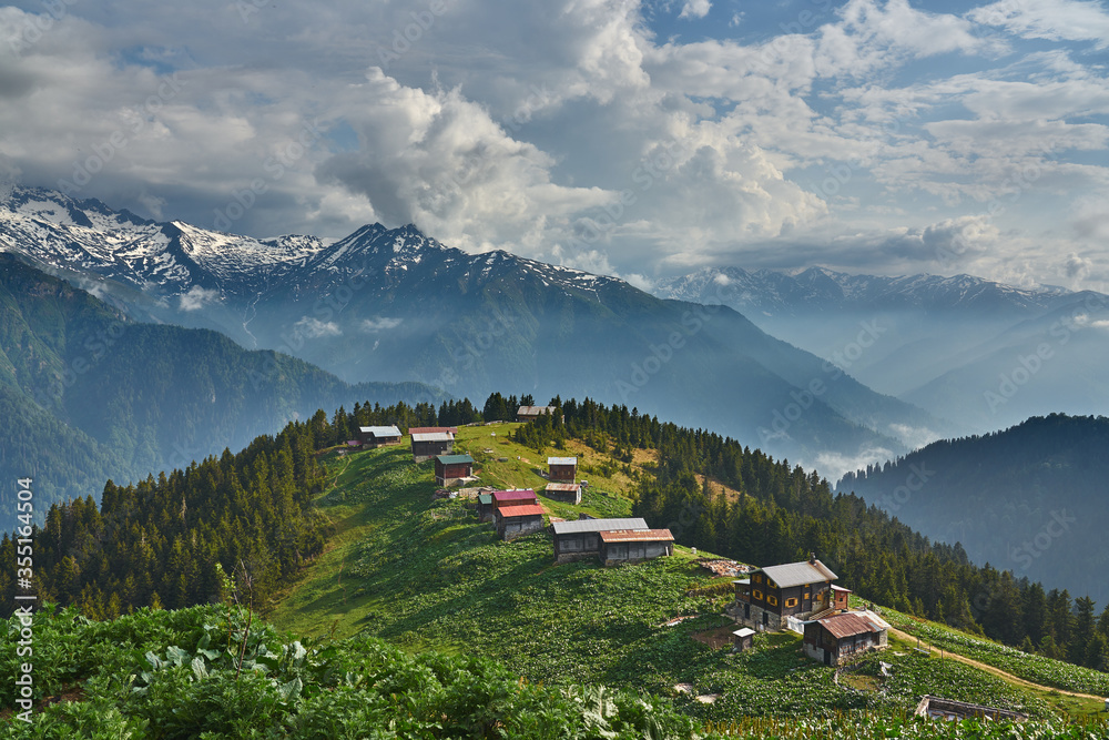 Landscape photo of Pokut Plateau with traditional wooden houses, snowy mountains, clouds and forest. Taken in summer at northeastern Black Sea / Karadeniz region of Turkey