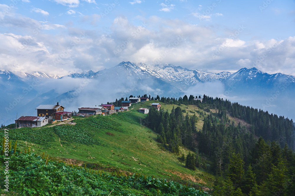 Landscape photo of Pokut Plateau with traditional wooden houses, snowy mountains, clouds and forest. Taken in summer at northeastern Black Sea / Karadeniz region of Turkey