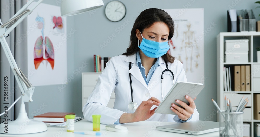 Caucasian young pretty female doctor in white gown and medical mask sitting at desk in cabinet and using tablet device. Woman medic scrolling, tapping and browsing on tablet computer online. Caucasian young pretty female doctor in white gown and medical mask sitting at desk in cabinet and using tablet device. Woman medic scrolling, tapping and browsing on tablet computer online.