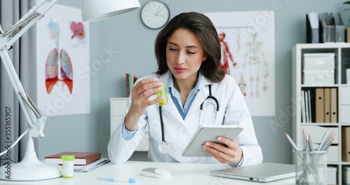 Beautiful Caucasian young female physician in white gown sitting at table in office and using tablet device with internet. Woman medic scrolling, tapping and browsing on tablet computer online.