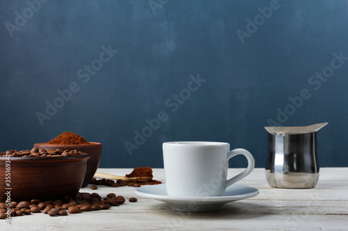 White espresso cup, Arabica beans in clay bowls, ground powder, metal Turkish pot on while table against dark blue wall. Side view, copy space. Coffee shop, morning, baristas workplace concept