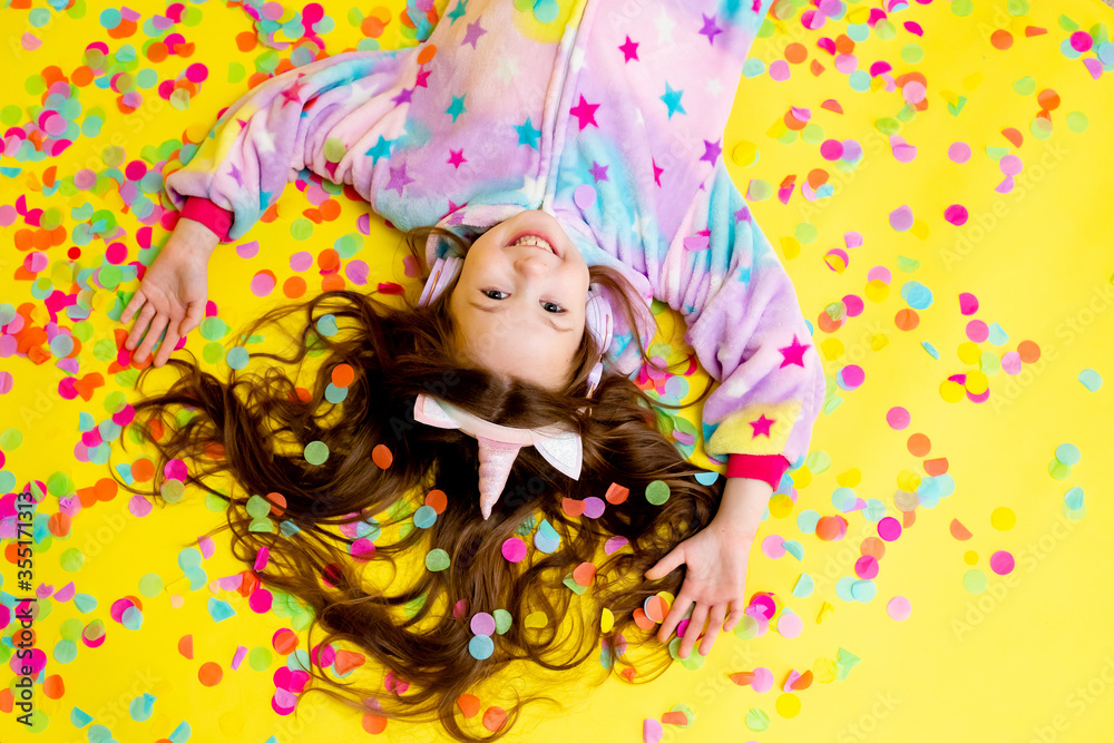 Fototapeta premium little girl with long hair wearing a kigurumi and headphones in the shape of a unicorn is happy catching confetti standing on a yellow background