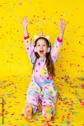 little girl with long hair wearing a kigurumi and headphones in the shape of a unicorn is happy catching confetti standing on a yellow background