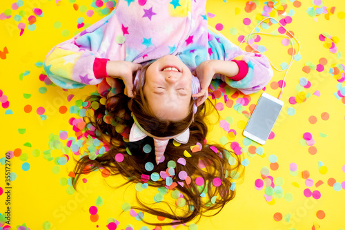 happy little girl in a unicorn kigurumi lies on a yellow background amid multicolored confetti listening to music