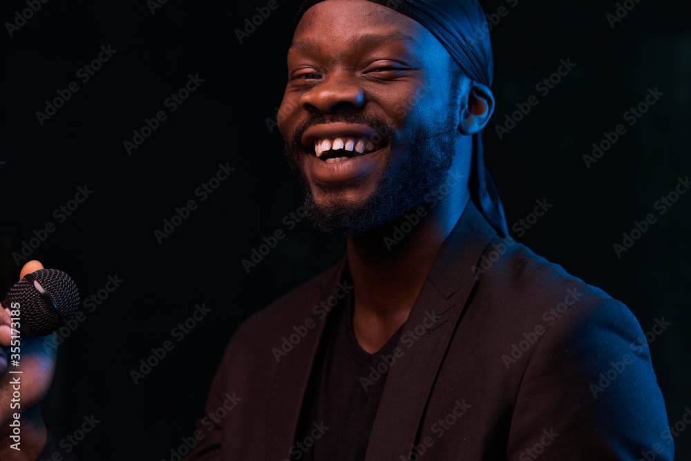 Fototapeta premium dark-skinned handsome guy in a bandana, black classic jacket and t-shirt holds a microphone in his hand, smiles and looks in the camera