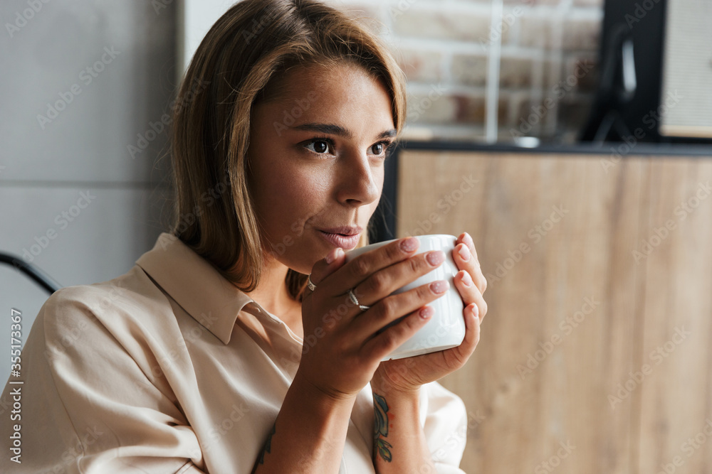 Image of young beautiful woman drinking tea while sitting on armchair