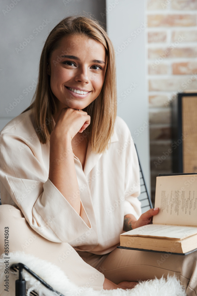 Image of young smiling woman reading book while sitting on armchair Image of young smiling woman reading book while sitting on armchair