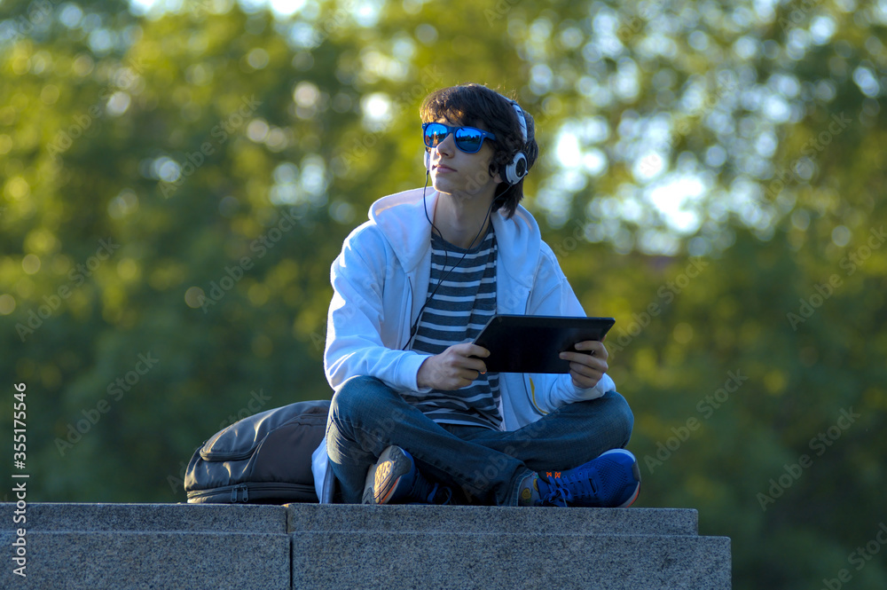 Obraz premium Young Man with Blue Sunglasses and Headphones, Sitting Cross-legged and Holding a Tablet