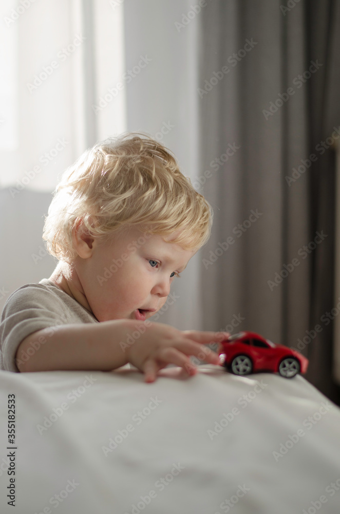 Blond toddler boy is playing with red toys car in bed. Children, play and development Blond toddler boy is playing with red toys car in bed. Children, play and development