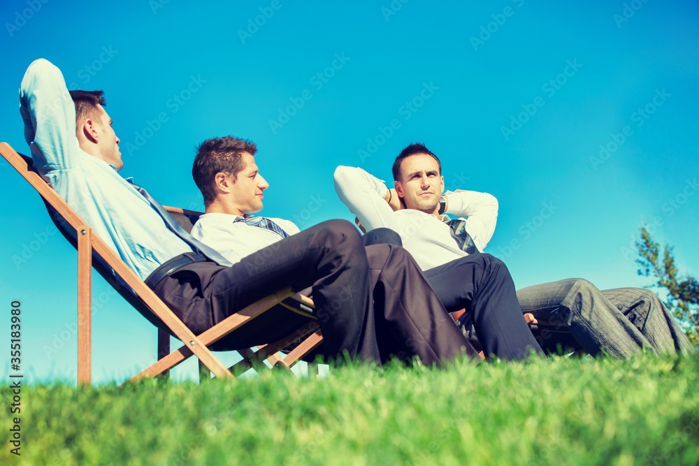 Group of handsome businessmen relaxing and talking about life while sitting on folding chair at park
