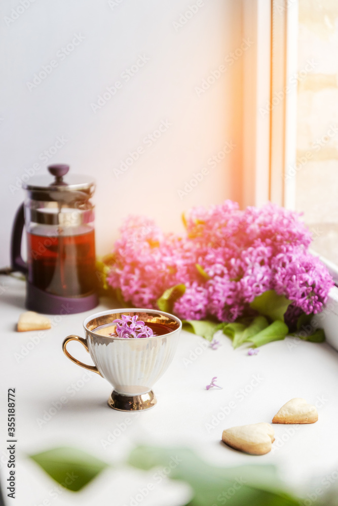 Fresh hot black tea in a beautiful porcelain cup with a lilac bouquet in the background on a white windowsill Fresh hot black tea in a beautiful porcelain cup with a lilac bouquet in the background on a white windowsill