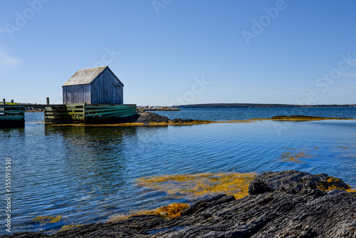 Lone wooden building on small rock outcropping in calm waters around Halifax, Nova Scotia with bright blue sky in background. 