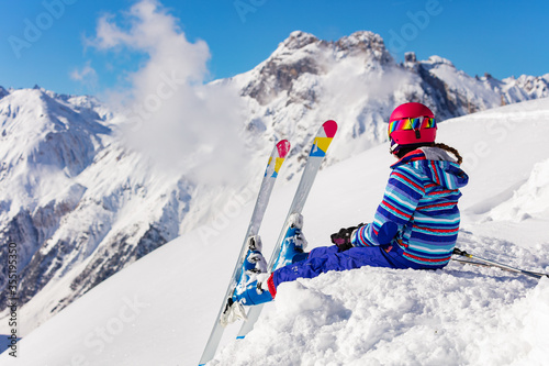 Happy young girl in bright sport outfit sit on the snow pile in the mountain over high peaks look on the valley view from behind